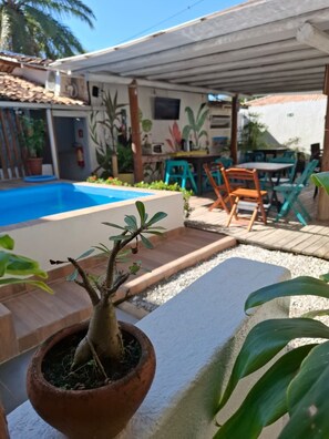 A pool area with a potted plant in the foreground, a covered patio with chairs, and a building with a tiled roof in the background. - Angatu (Porto Seguro)
