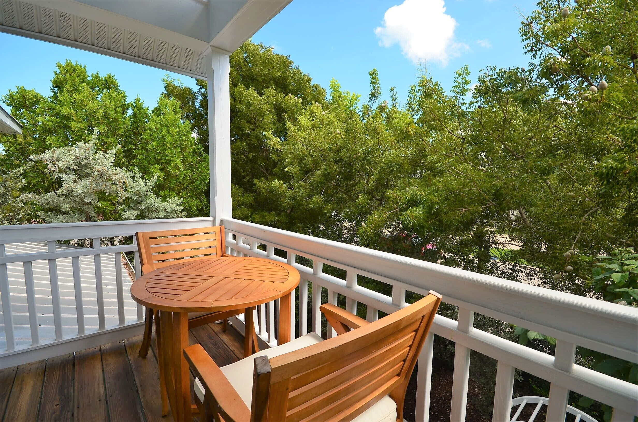 Une terrasse en bois avec une table ronde et des chaises, entourée de rampes blanches, offrant une vue sur un paysage verdoyant et luxuriant.