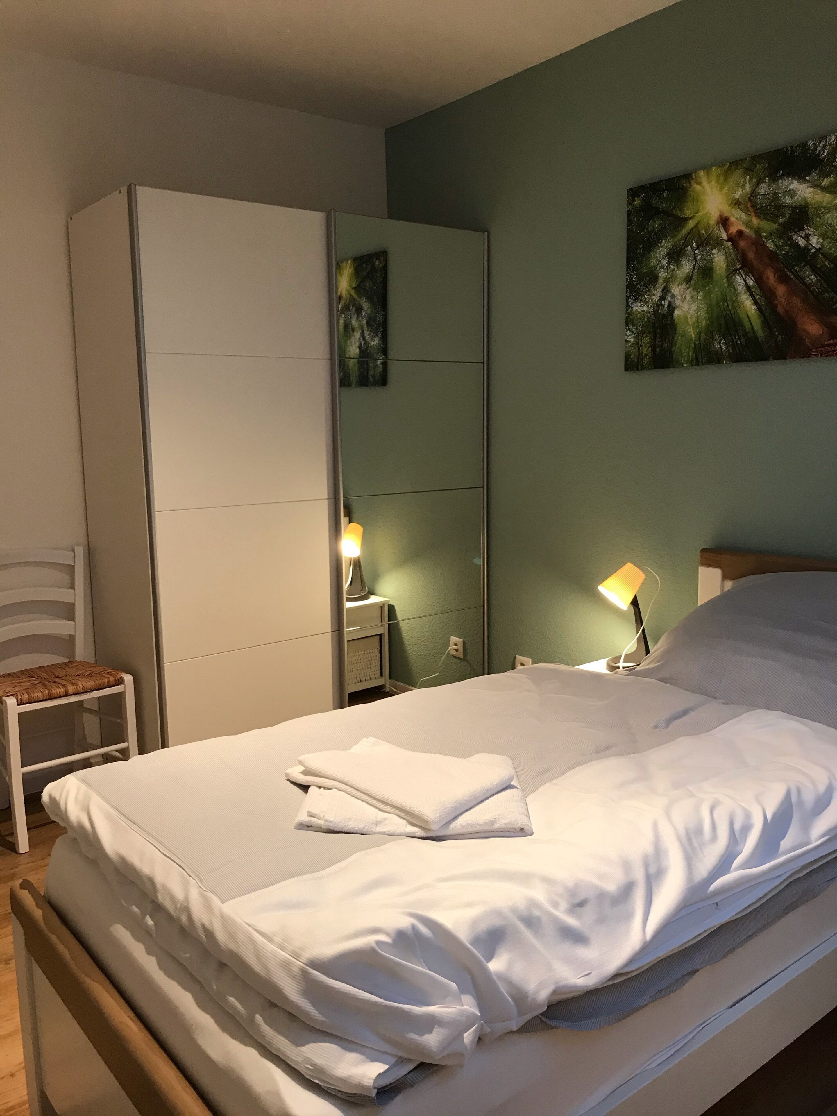 Ein ordentlich bezogenes Bett mit weißen Leinen, ein Nachttisch mit Lampe, ein Sessel und ein Kleiderschrank.