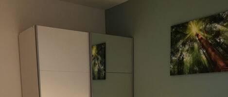 Ein ordentlich bezogenes Bett mit weißen Leinen, ein Nachttisch mit Lampe, ein Sessel und ein Kleiderschrank.