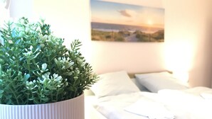 A neatly made bed with white bedding, a white pillow, and a potted plant with green foliage on a bedside table. - Luxus Ferienwohnung für 6 P. mit Sauna & Holzofen - am Wasser und in Strandnähe (Geeste)