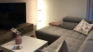 A modern living room with a grey sofa, a flat-screen TV, a white coffee table, and a vase with flowers. - Luxus Ferienwohnung für 6 P. mit Sauna & Holzofen - am Wasser und in Strandnähe (Geeste)