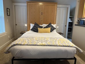 A neatly made bed with a floral-patterned bedspread, two nightstands with lamps, a wooden wardrobe, and a closet with shelves. - Hillside Loft- Spacious Studio, 100+mbps Wifi, Gas Fireplace, AC, Balcony (Bend)
