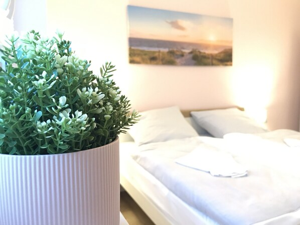 Ein ordentlich bezogenes Bett mit weißer Bettwäsche, einem weißen Kopfkissen und einem Topf mit Grünpflanzen auf einem Nachttisch.