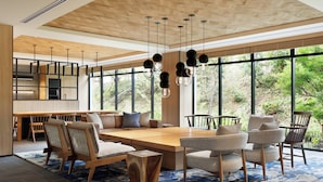 Un vaste espace dédié à la salle à manger, avec une table et des chaises en bois, de grandes fenêtres donnant sur un paysage verdoyant, et des suspensions lumineuses dotées d’abat-jours sphériques.