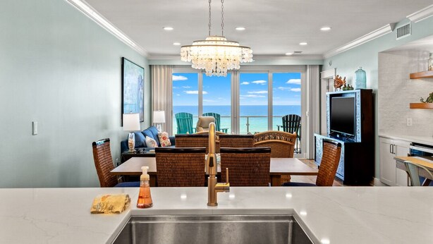 A modern kitchen with a large island, dining area with wicker furniture, a television, and a view of the ocean. - Sterling Sands 313 - On the Beach! Renovated 2B/2B w/Pool/Hot Tub/Garage Parking (Destin)