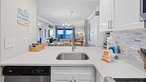 A modern kitchen with white cabinetry, a built-in dishwasher, and a large sink. There is a wicker basket on the counter and a bag of kitchen towels.