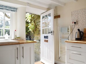 Une cuisine avec des meubles blancs, un plan de travail en bois, une vue sur la verdure à l’extérieur, une bouilloire et une prise électrique murale.