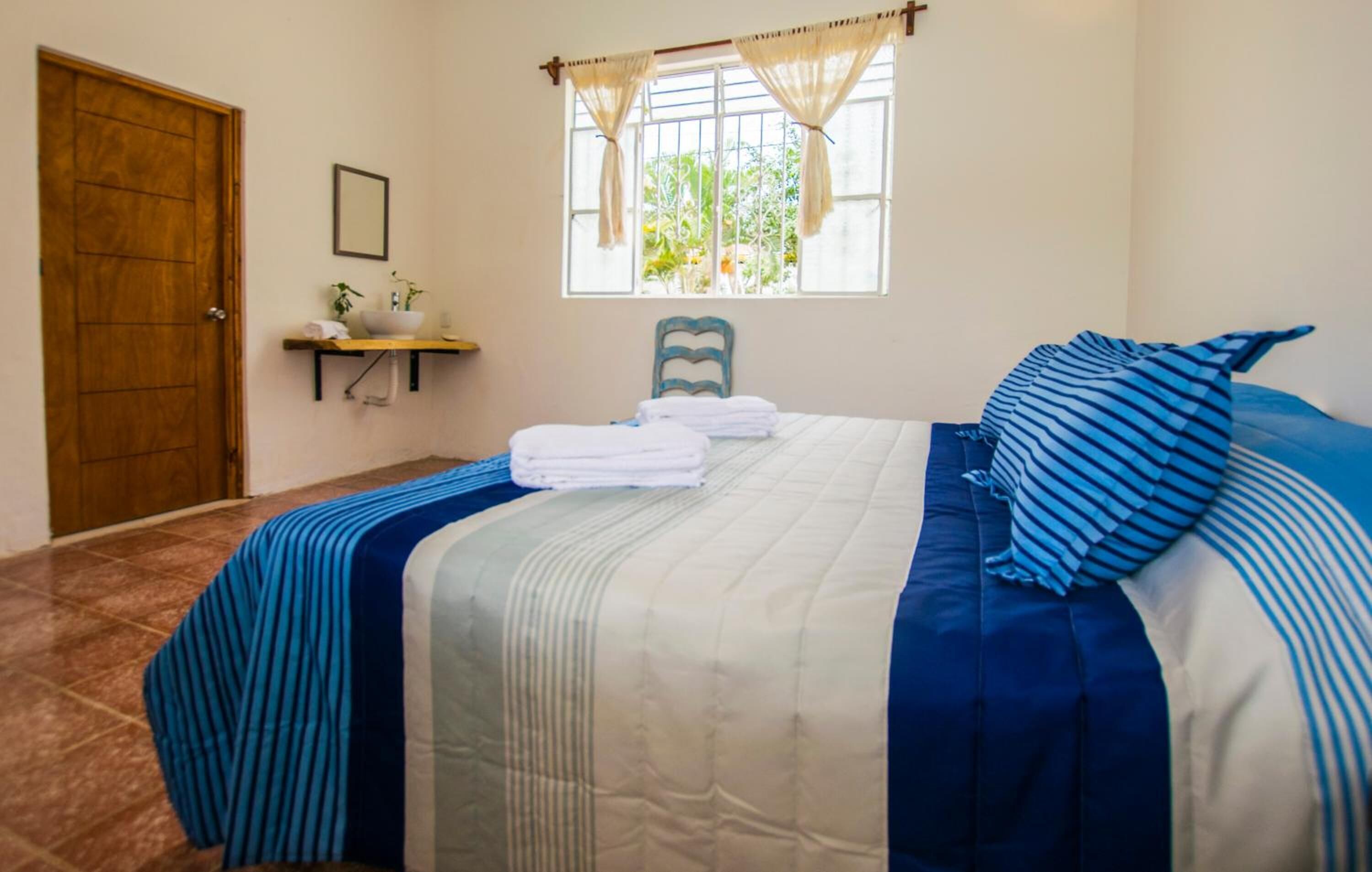 Une chambre à coucher avec un lit, une fenêtre avec des rideaux, une porte en bois, une petite étagère avec une plante et une chaise.