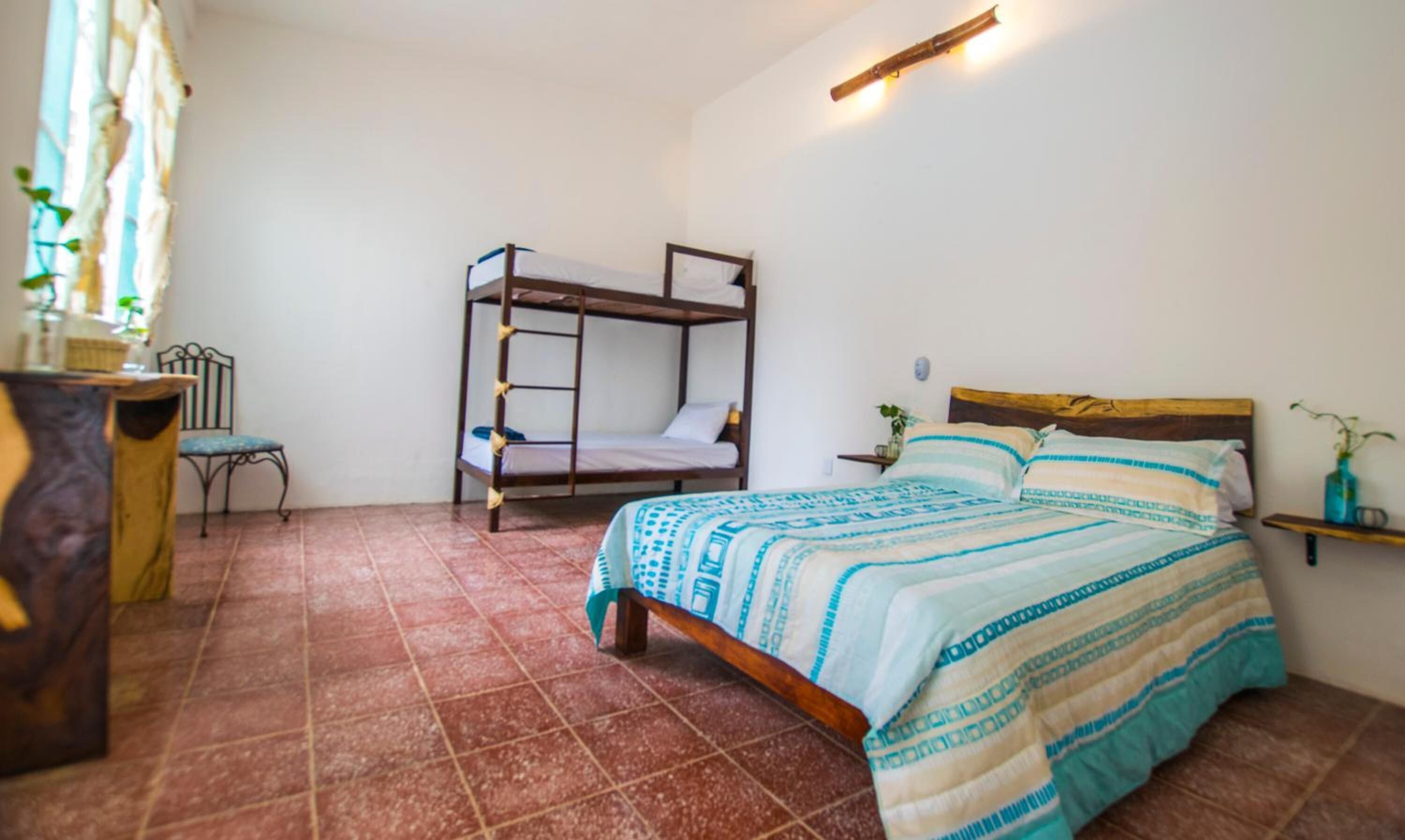 A room with a bunk bed, a single bed, a small table, a chair, and a shelf with a plant.