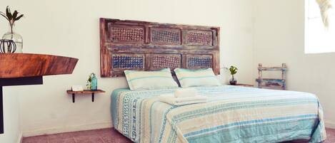 Un dormitorio con una cama, cabecera, estante con planta y una mesita con planta.