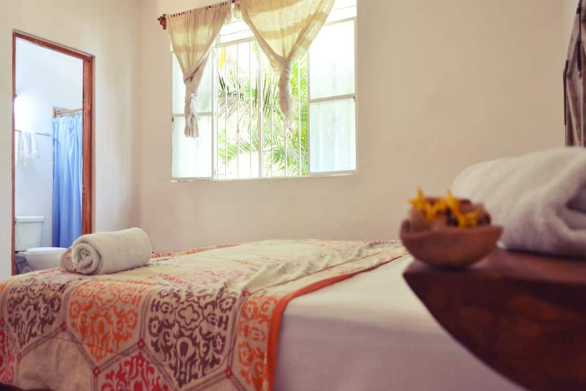 Una habitación con una cama, una ventana con cortinas, una toalla sobre la cama y una mesa de madera con un cuenco.