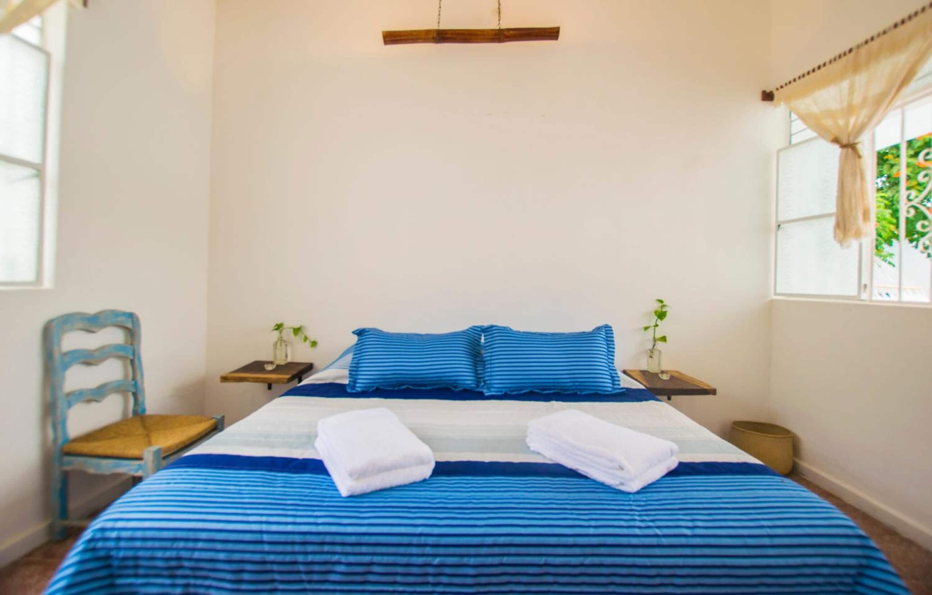 A neatly made bed with blue bedding, two bedside tables with lamps, a chair, and a window with curtains.