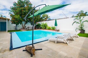 Una piscina con un paraguas verde, sillas de descanso blancas y diversas plantas.