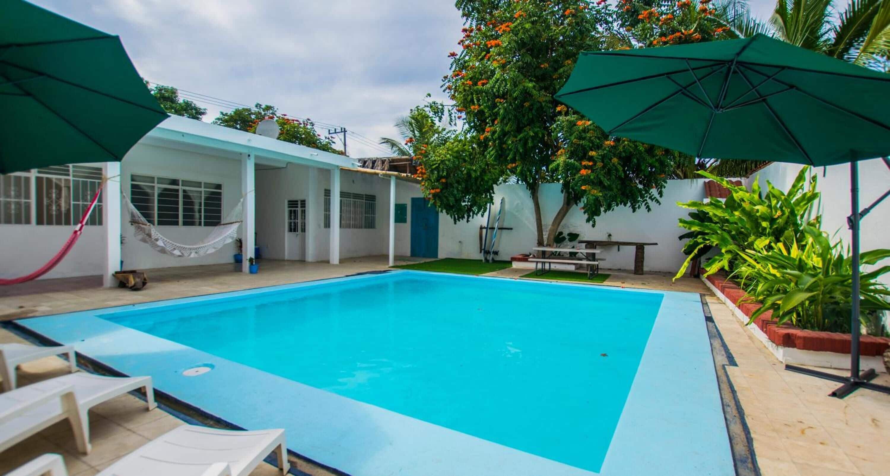 Una piscina rectangular amb interior blau, envoltada per parets blanques i una zona de pati coberta amb ombrelles verdes.