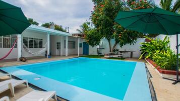 Una piscina rectangular con interior azul, rodeada de paredes blancas y un área de patio techada con sombrillas verdes.