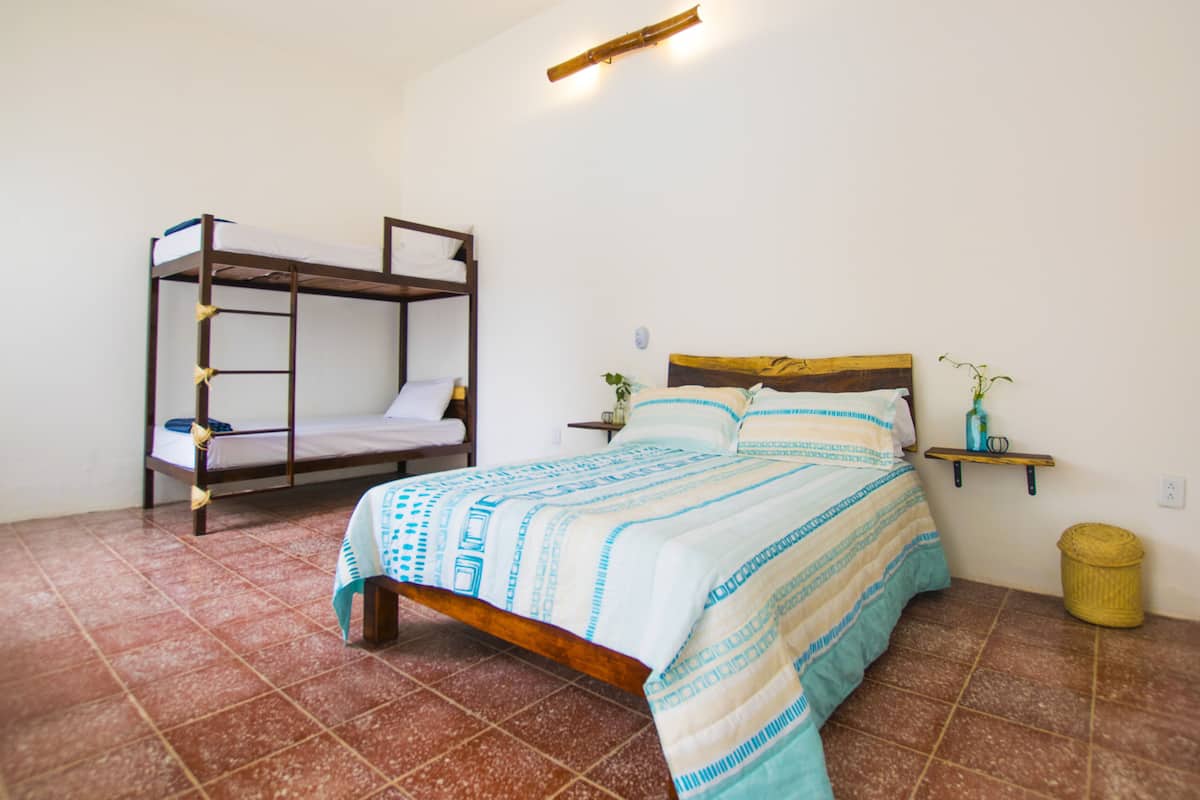 Una habitación con una litera, una cama individual, una mesita de noche con lámpara, un pequeño estante con una planta y un cubo de basura.