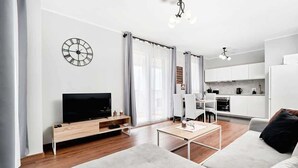 A modern living room with a flat-screen TV, a sofa, a coffee table, and a dining area with a kitchen in the background.