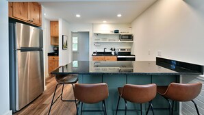A modern kitchen with a stainless steel refrigerator, a microwave, and a stove. There are three bar stools and a large island with a black countertop. - All 4 lakefront condos! Private beach, dock, sauna, club room, decks. (Webb Lake)