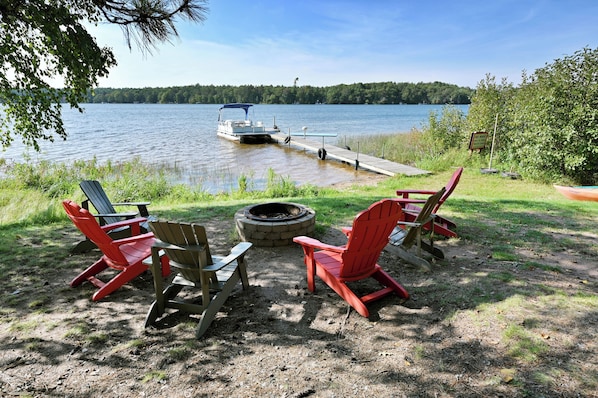 Une terrasse au bord du lac, avec des fauteuils Adirondack, un foyer et un quai avec un bateau.