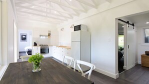 A modern kitchen with white cabinetry, a built-in oven, and a refrigerator. There is a dining table with chairs and a vase of green plants. - Maple Cottage - Gorgeous character filled cottage (KANGAROO VALLEY)