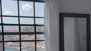 A hotel room with a large window offering a view of a harbor and buildings. There is a bed with a quilted cover and a dark-colored dresser with drawers. - Ocean View (St, George)
