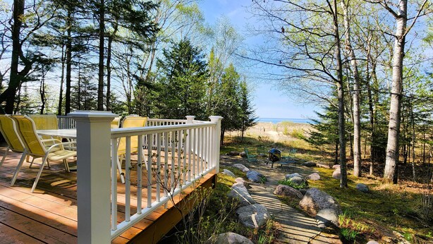 Une terrasse en bois, agrémentée de mobilier d’extérieur, une balustrade blanche et une vue sur une zone boisée sous un ciel dégagé.