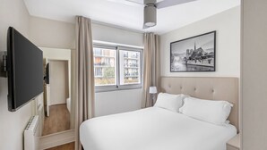 A hotel room with a large bed, a flat-screen TV, a window with curtains, a ceiling fan, and a cityscape painting. - Large 2-Bed Apartment | Paris Vincennes | Sleeps 6 (Paris)