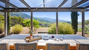 A covered outdoor dining area with a table set for two, overlooking a vineyard and hilly landscape.