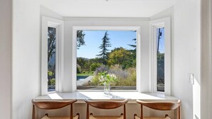 A dining area with a table and four chairs, a vase with flowers, and a view of a garden and trees through large windows.
