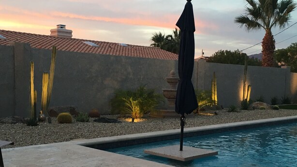 A swimming pool with a tiled deck, surrounded by a landscaped garden with cacti and palm trees, and a sunset in the background. - Beautiful Backyard Oasis Lake House (Lake Havasu City)