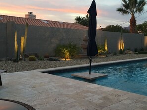 A swimming pool with a tiled deck, surrounded by a landscaped garden with cacti and palm trees, and a sunset in the background. - Beautiful Backyard Oasis Lake House (Lake Havasu City)
