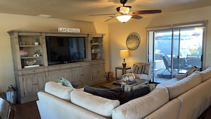 A living room with a large TV, a sofa, a coffee table, a lamp, and a ceiling fan. - Beautiful Backyard Oasis Lake House (Lake Havasu City)