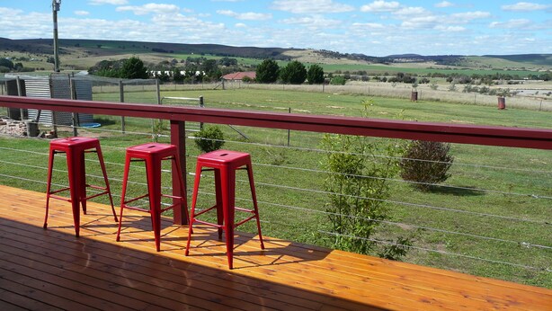 A wooden deck with red barstools and a metal railing overlooking a grassy field with distant hills.
