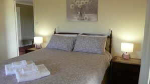 A neatly made bed with a floral-patterned bedspread, two pillows, and folded towels on the bed. There is a painting of flowers on the wall above the bed.