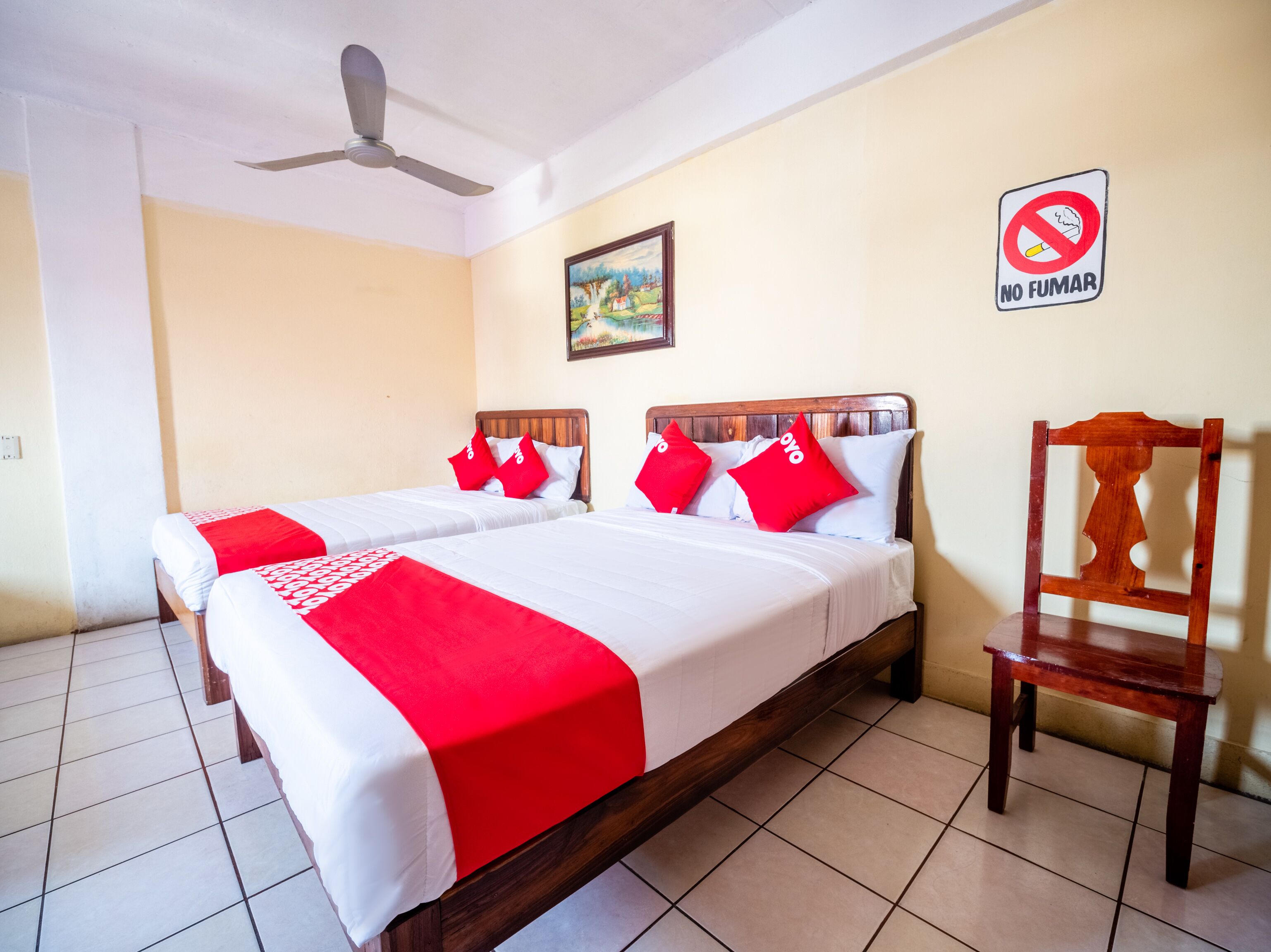 A hotel room with two beds, a wooden chair, a ceiling fan, a painting on the wall, and a 'No Smoking' sign.