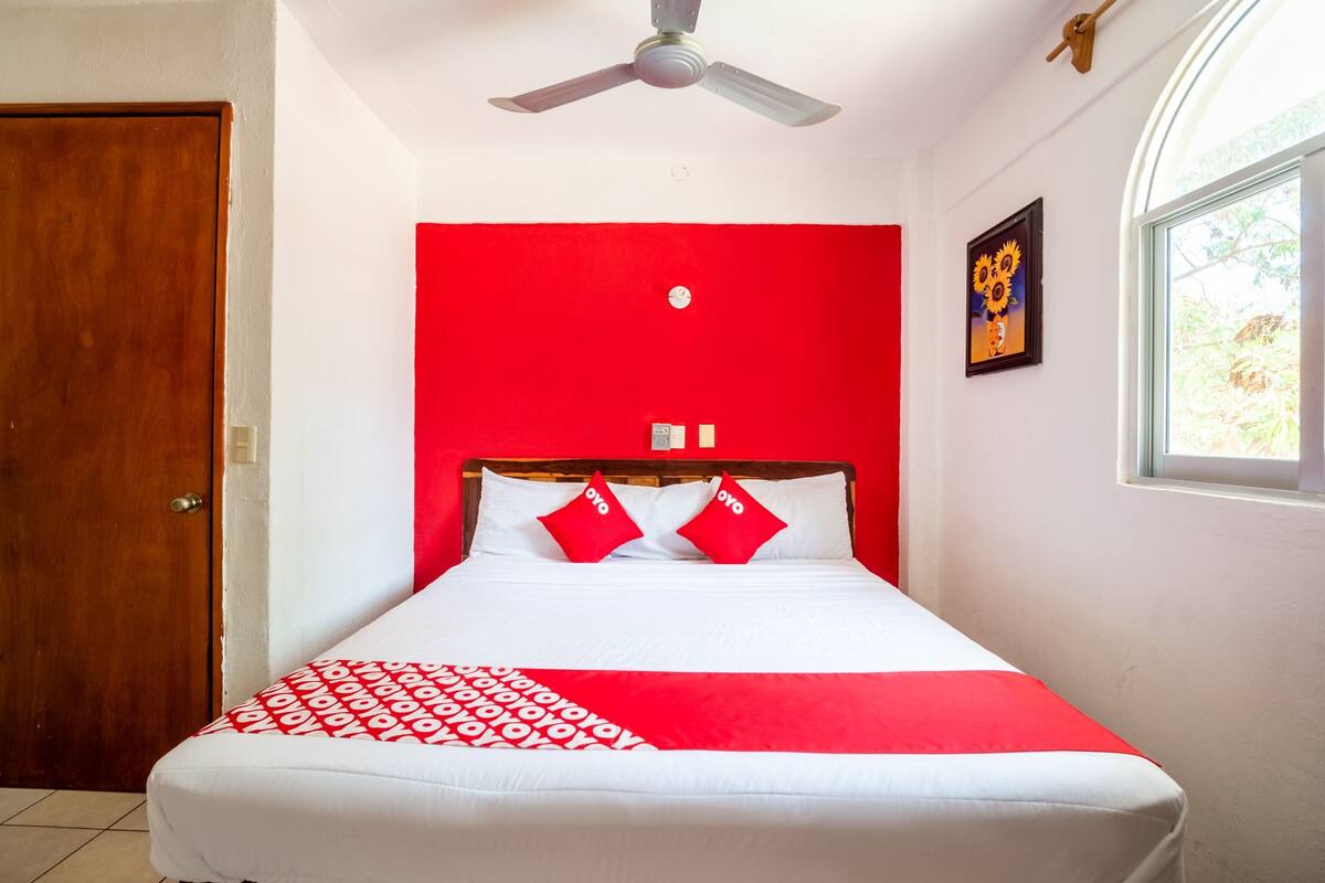 Ein Bett mit roter und weißer Bettdecke, ein Deckenventilator, eine Holztür und ein Fenster mit Blick auf Grünfläche.