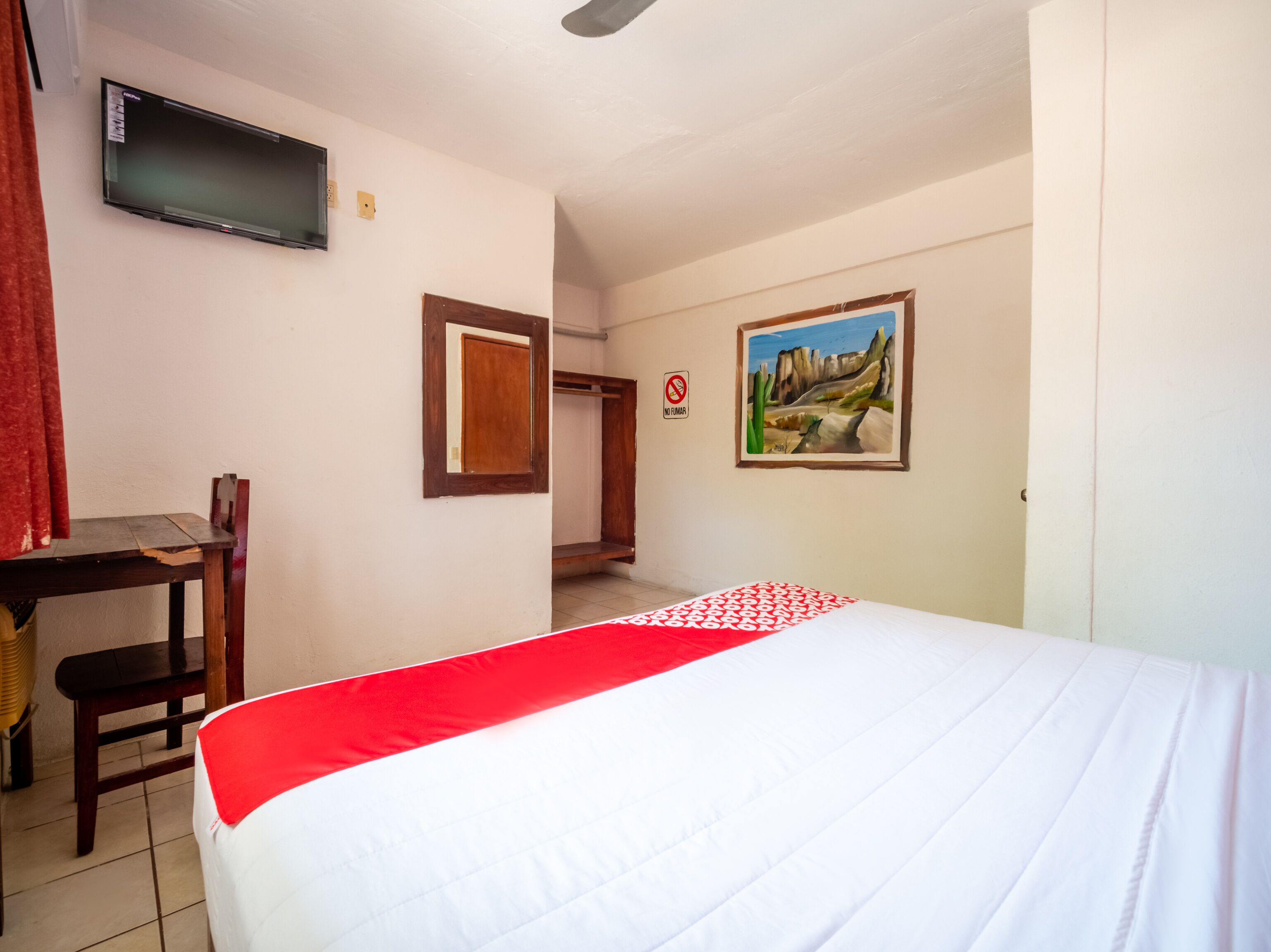 Habitación de hotel con cama, escritorio, silla, televisión, un cuadro y una puerta.