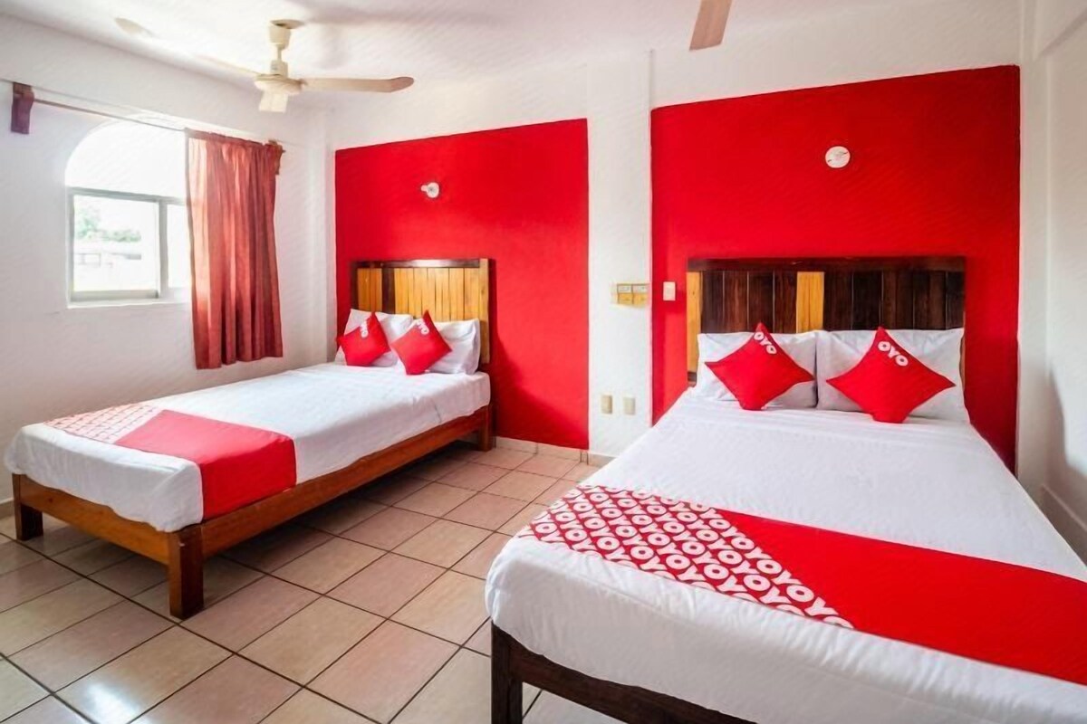 Ein Hotelzimmer mit zwei Betten, roten Akzenten, einem fensterbelaubten Fenster und einem Deckenventilator.