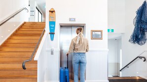 A woman with a suitcase approaching an elevator in a hotel lobby with wooden flooring and a skateboard on the wall. - The Surf House - Hostel (Byron Bay)