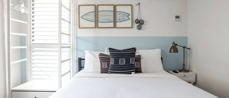 Un dormitorio con cama, mesita de noche, lámpara, aire acondicionado y ventana con persianas.