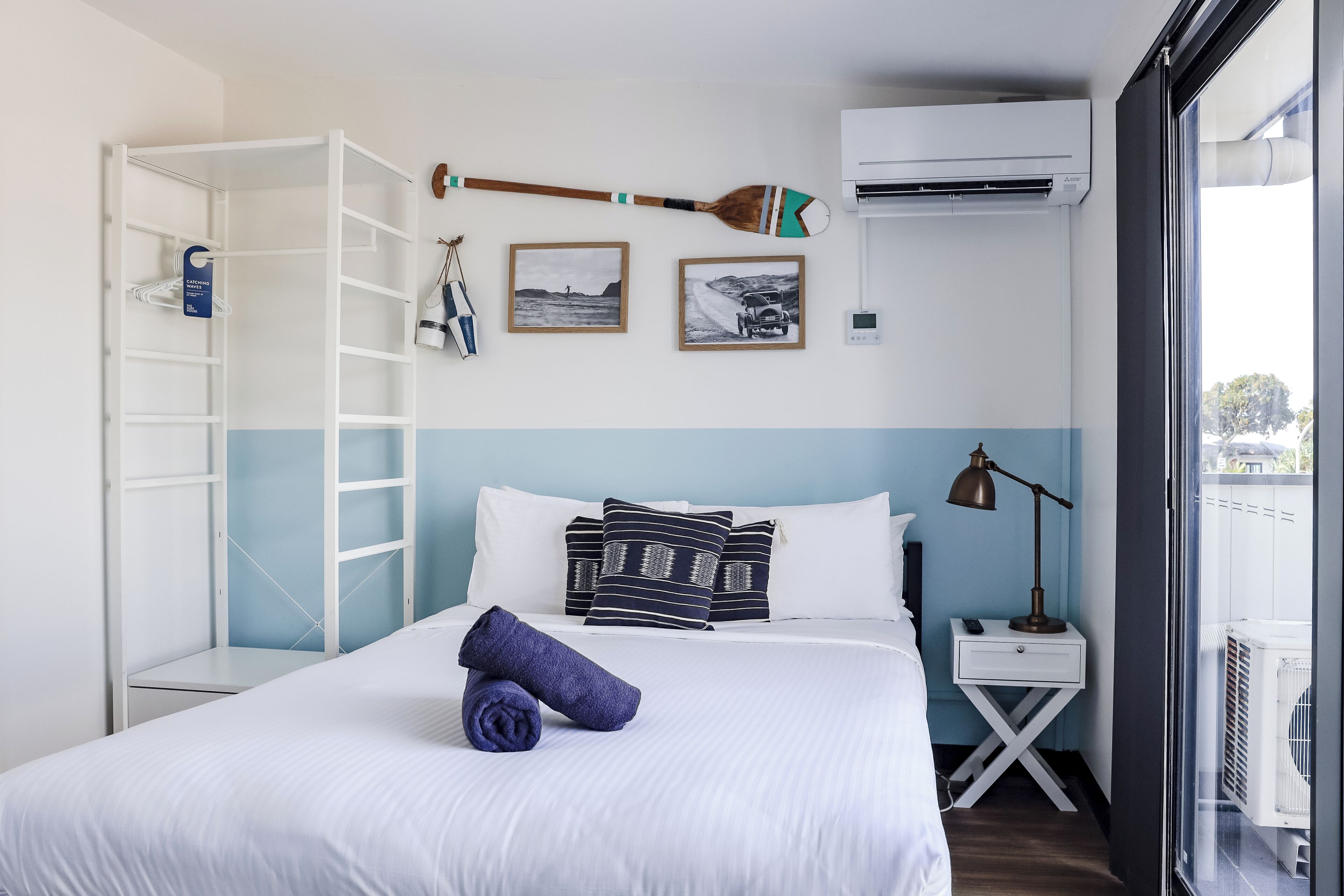 A hotel room with a bed, a ladder, a wall-mounted air conditioner, a small table with a lamp, and a window with a view.