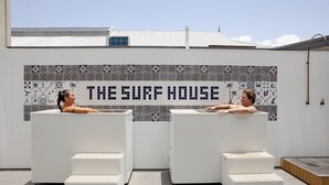 Two white lounge chairs with a tiled wall behind them, featuring a pattern of blue and white tiles and the text "THE SURF HOUSE". - The Surf House - Hostel (Byron Bay)