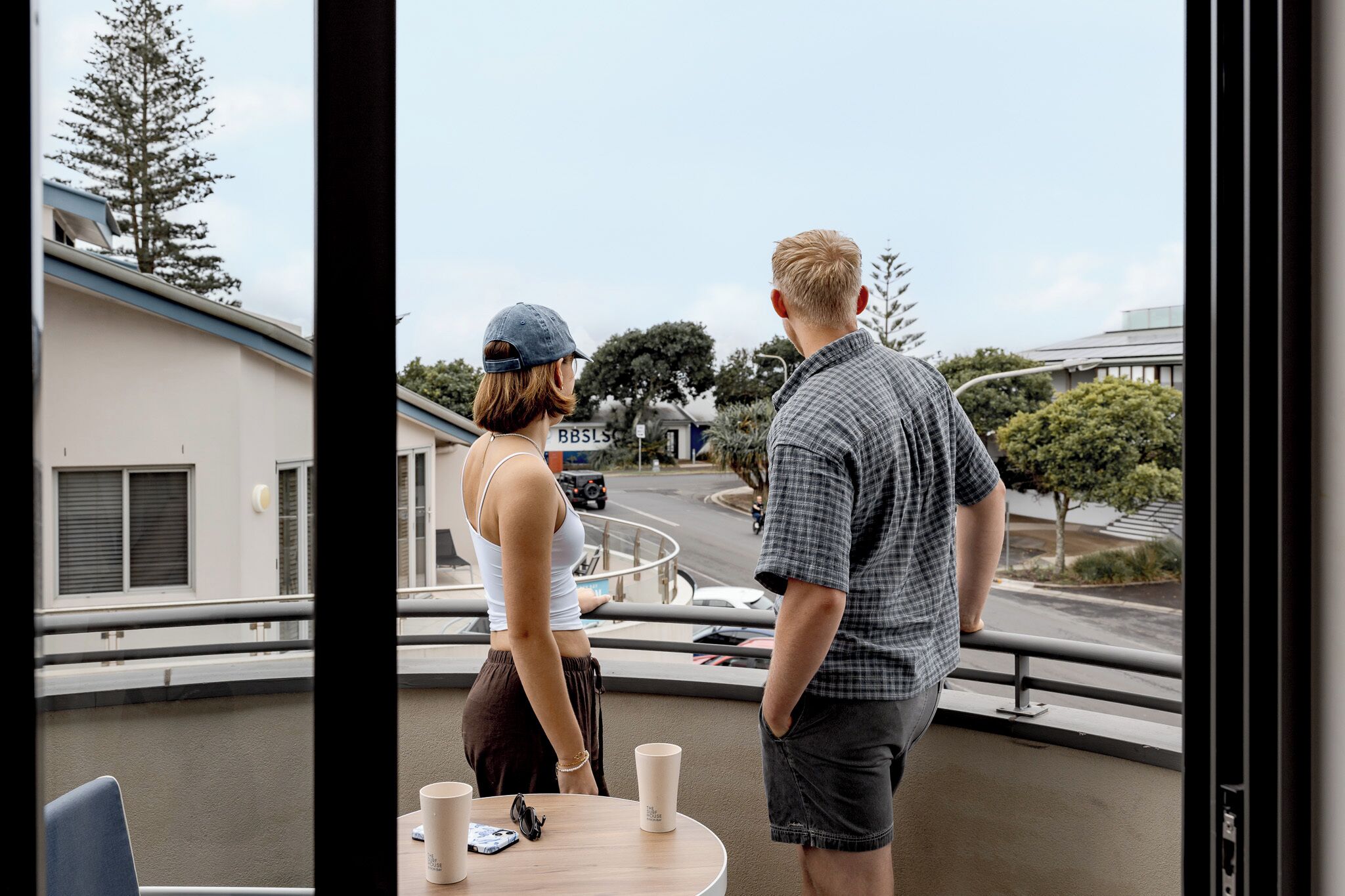 A couple standing by a window overlooking a street with buildings and trees.