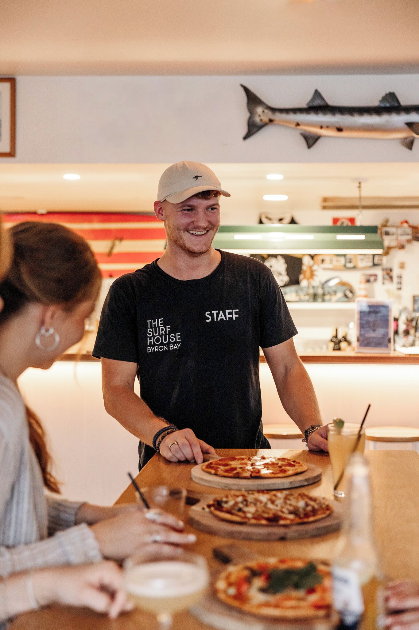 A person wearing a black t-shirt with 'STAFF' and 'THE SURF HOUSE BYRON BAY' printed on it, standing behind a counter with pizza and a drink.