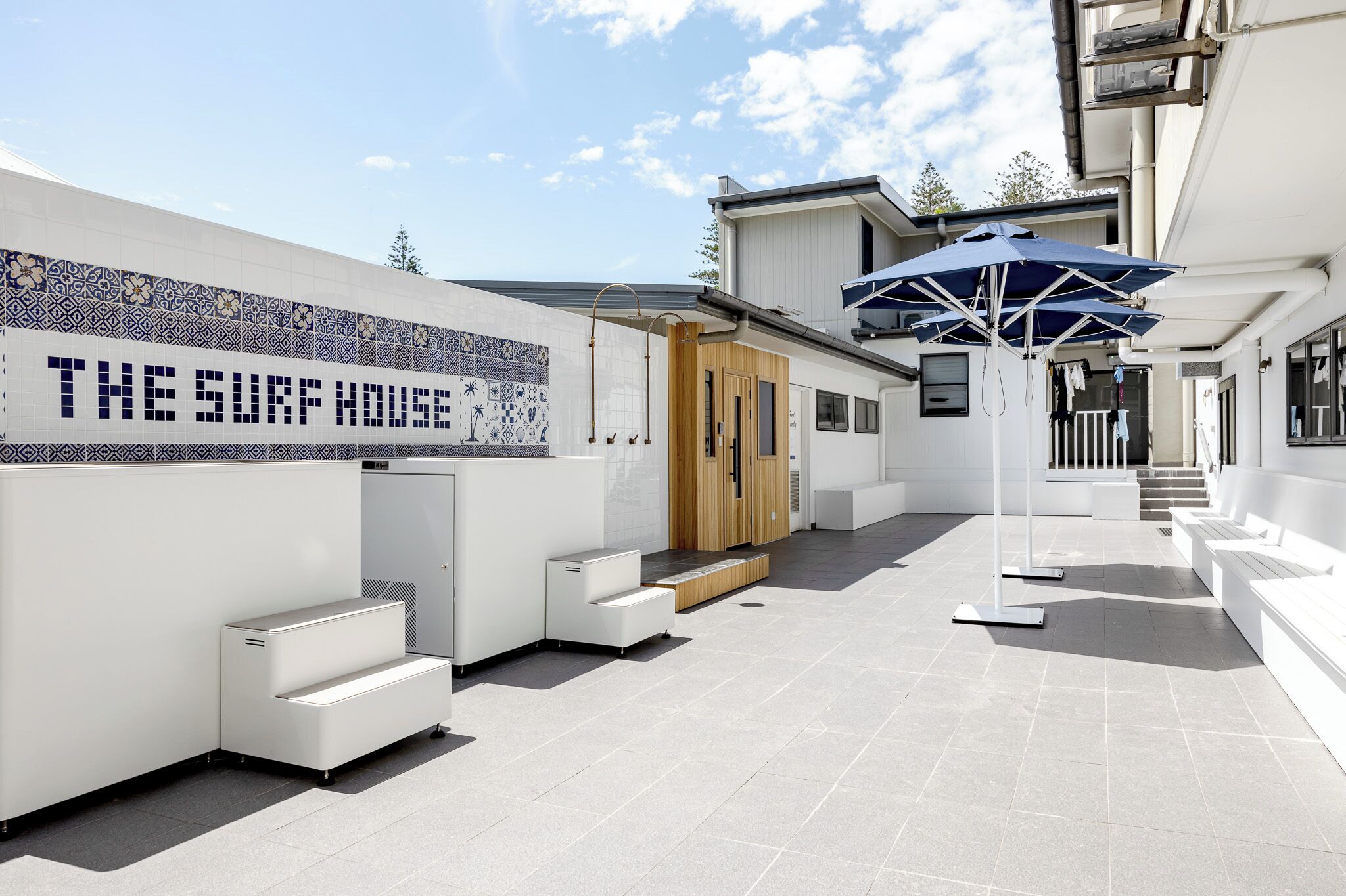 A modern building with a tiled wall reading "THE SURF HOUSE" and a covered seating area with steps leading to a wooden door.