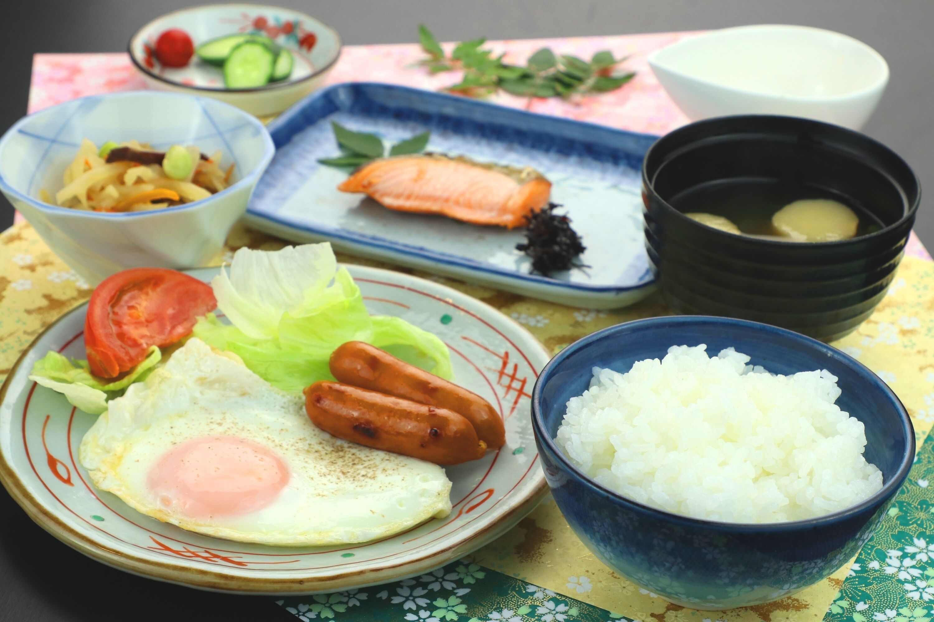 A table with various dishes including a plate with an egg, sausages, and vegetables, a bowl of rice, and a plate with salmon.