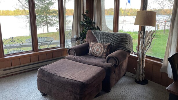 Ein gemütliches Wohnzimmer mit einem braunen Sofa und Hocker, großen Fenstern mit Vorhängen, einer Lampe und Blick auf einen See und Bäume.