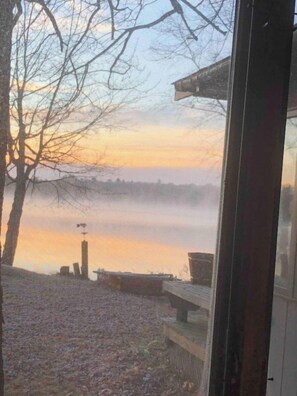 Blick durch ein Fenster auf ein Holzdeck über ruhigem Wasser, mit kahl stehenden Bäumen und weiter Ferne.