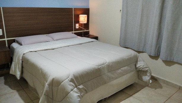 A neatly made bed with a white comforter and pillows, a wooden headboard, a bedside table with a lamp, and a curtain on the window. - 2 bedroom house near UNESP (Botucatu)
